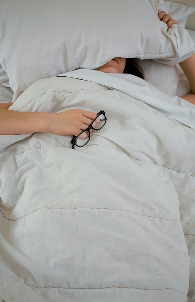 person in bed with face partially covered by a pillow, holding a pair of spectacles